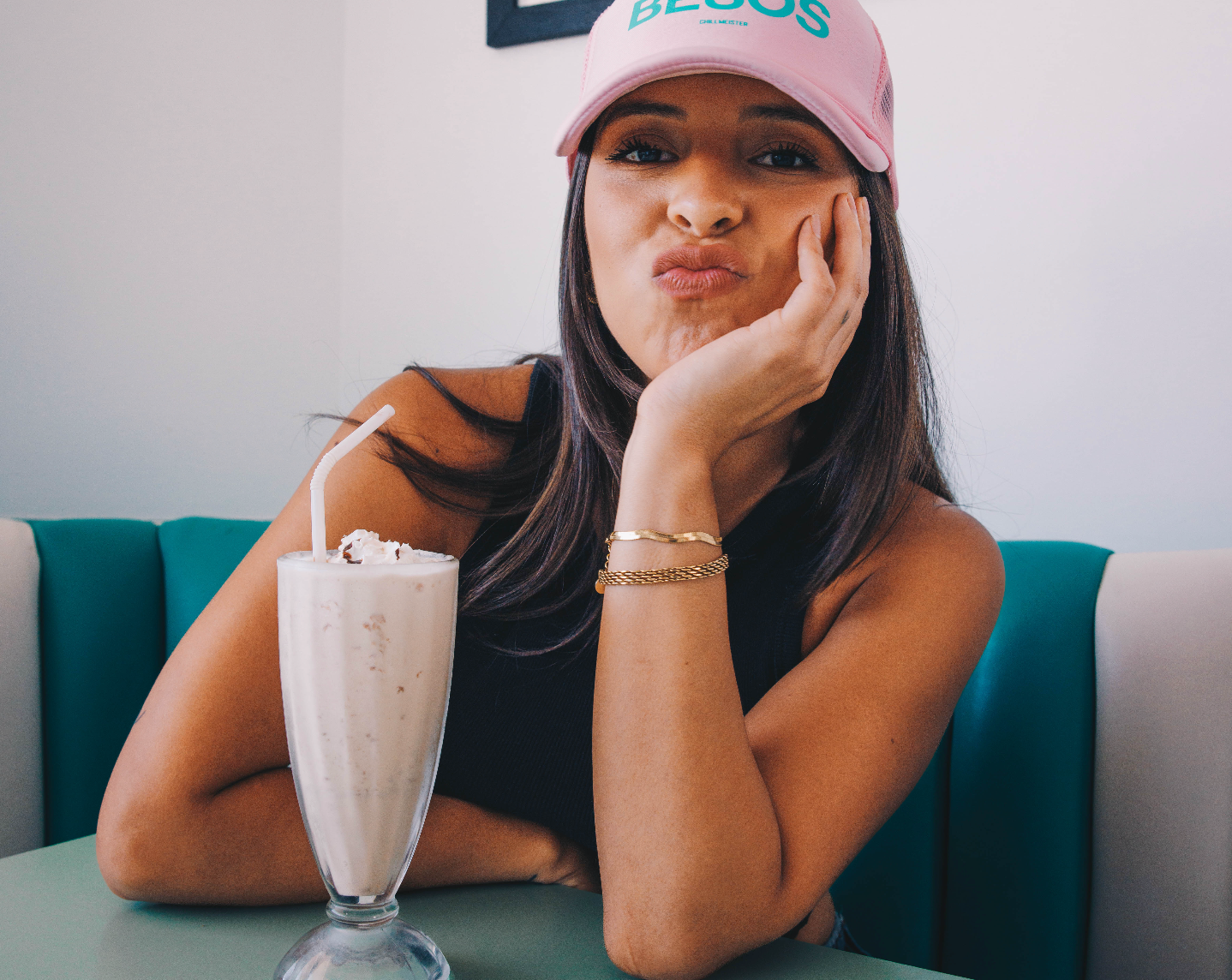 A young woman in a pink cap sits at a table with a milkshake, resting her chin on her hand, with a playful expression.